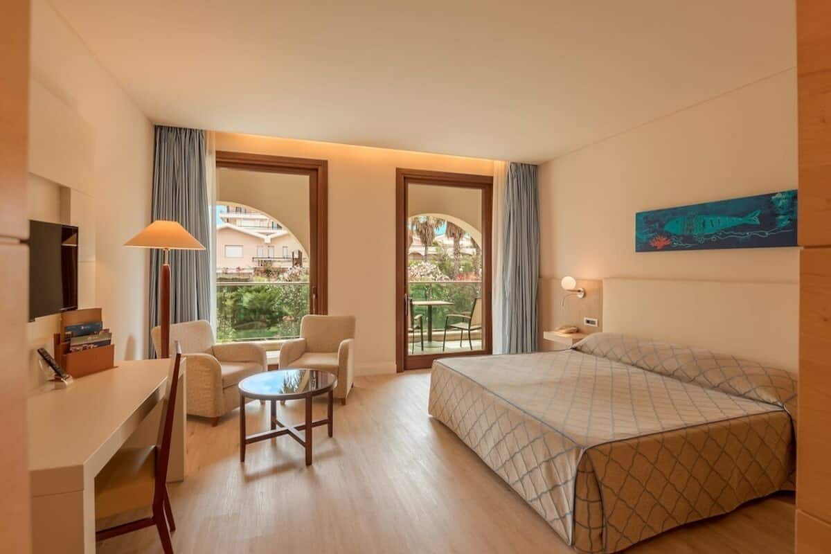 A hotel room with a bed, desk, chair, and a small table. There is a balcony with a view of the outside.