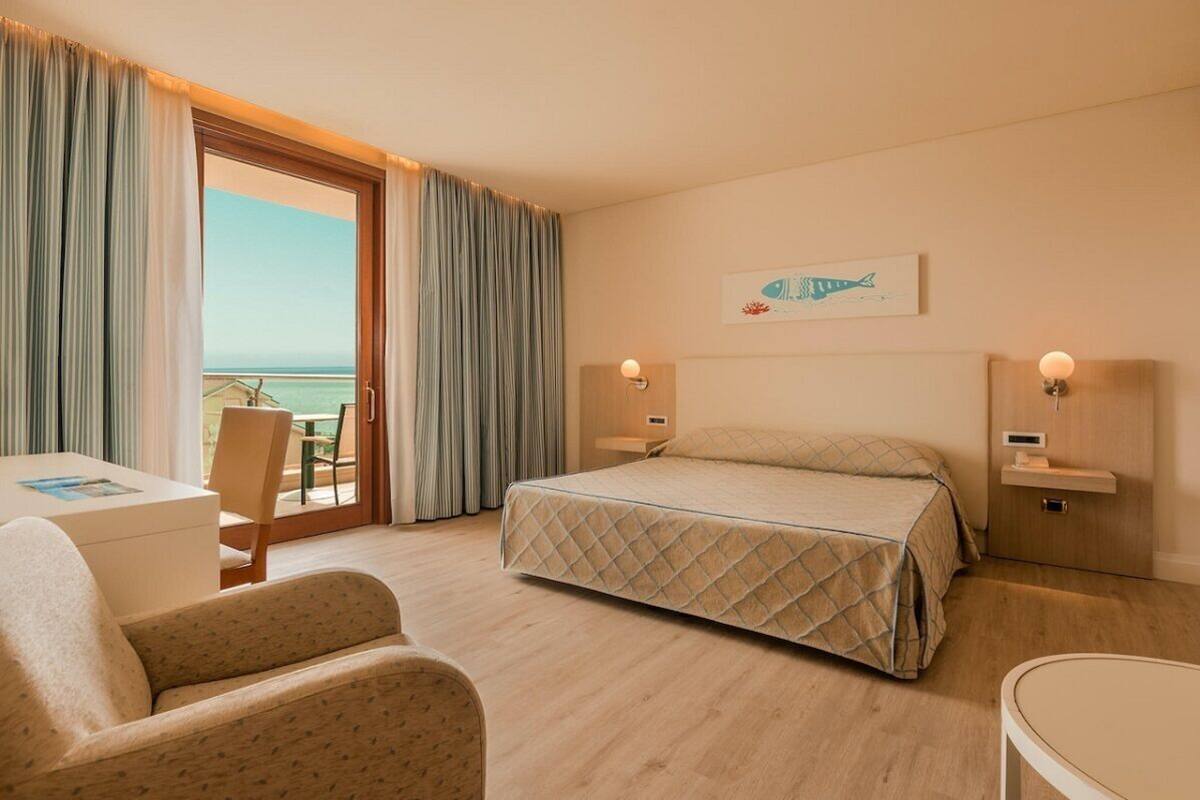 A hotel room with a bed, a desk, a chair, a small table, and a view of the ocean.