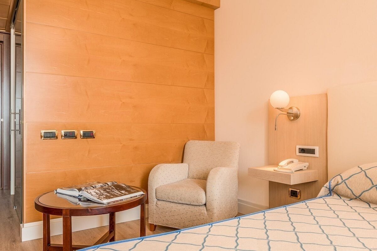 A hotel room with a wooden panel wall, a small round table with a magazine, a beige armchair, a bedside table with a lamp, and a bed with a patterned bedspread.