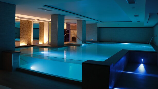 A modern indoor swimming pool with clear water, surrounded by walls with patterned tiles and a few pillars. - Smy Carlos V Alghero (Alghero)