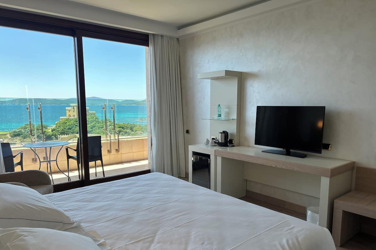 A hotel room with a large bed, a desk with a television, and a view of the sea.