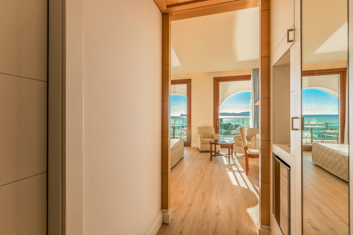 A modern hotel room with a wooden floor, large windows offering a view of the city, a bed, and a seating area with a table and chairs.