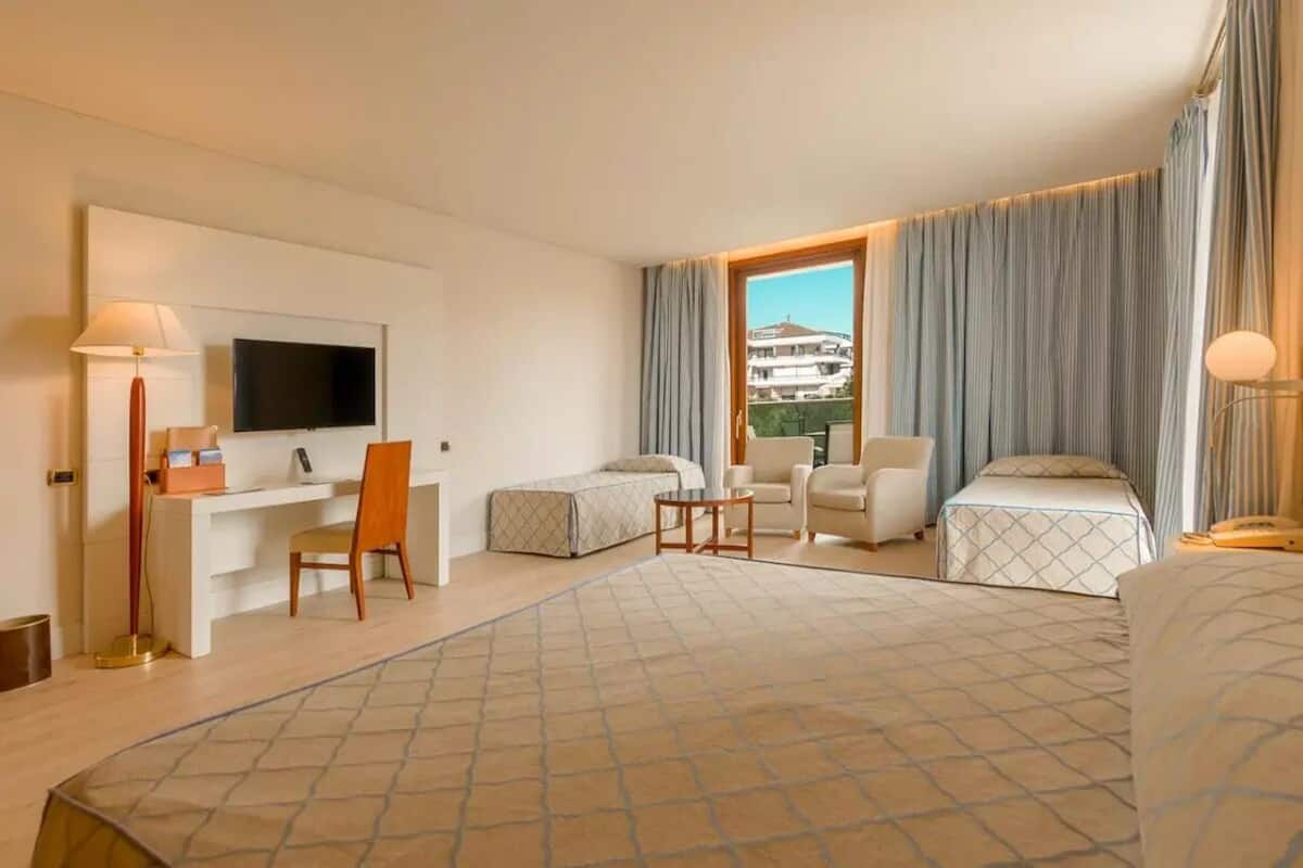 A hotel room with a bed, two chairs, a desk with a TV, and a window with curtains.
