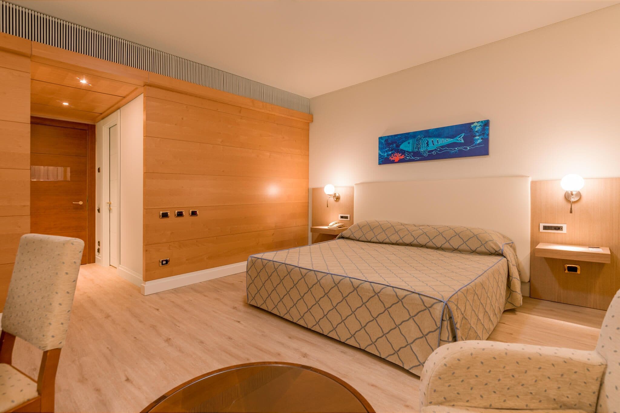 Een moderne slaapkamer met een houten vloer, een bed met een deken met een geometrisch patroon, twee nachtkastjes met lampen, een kunstwerk aan de muur, en een deur naar een andere kamer.