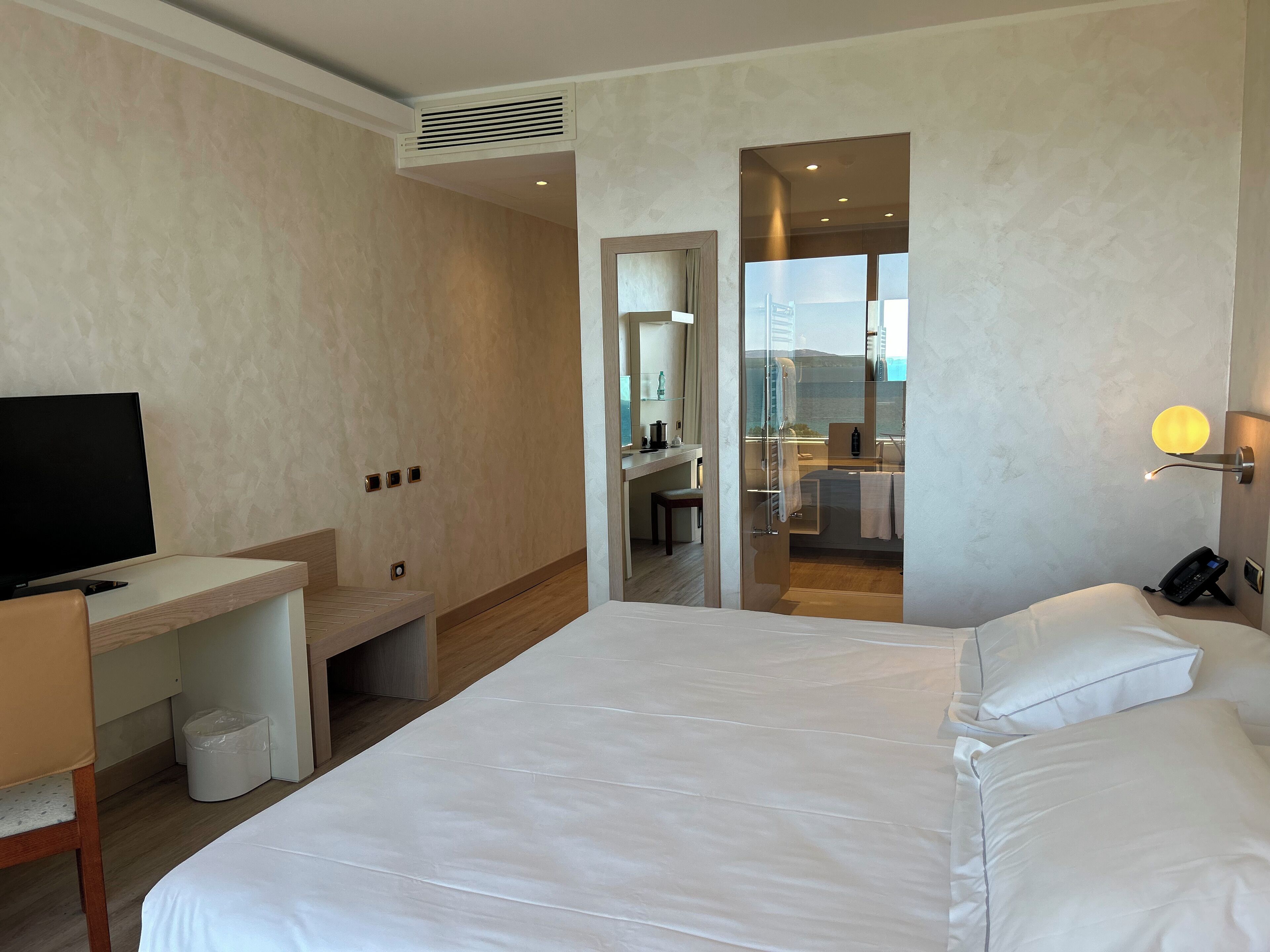 A hotel room with a bed, a desk, a TV, and a bathroom visible through a glass door.