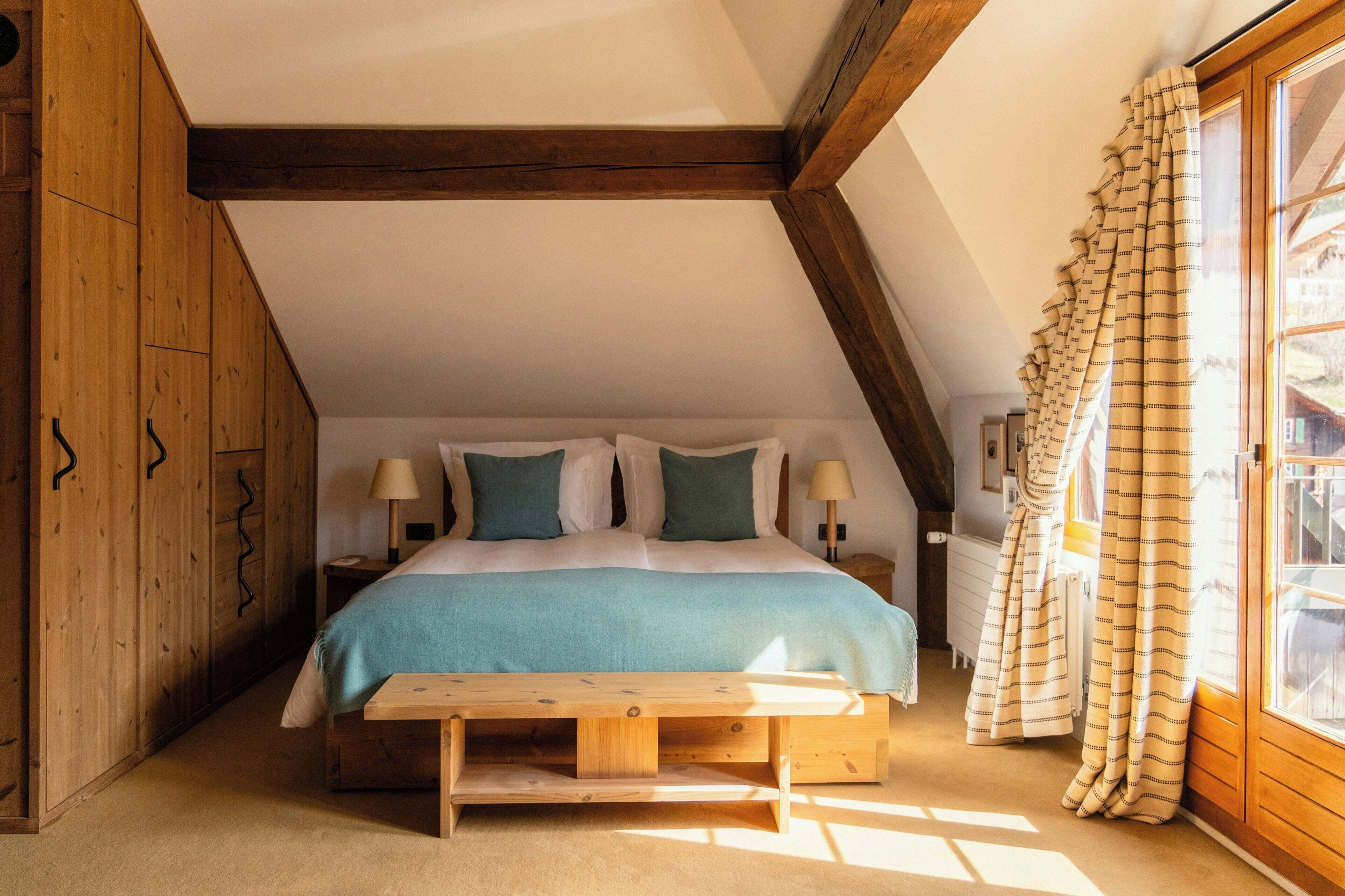 A bedroom with a wooden ceiling, a bed with white and teal bedding, a bedside table with lamps, and a window with curtains.