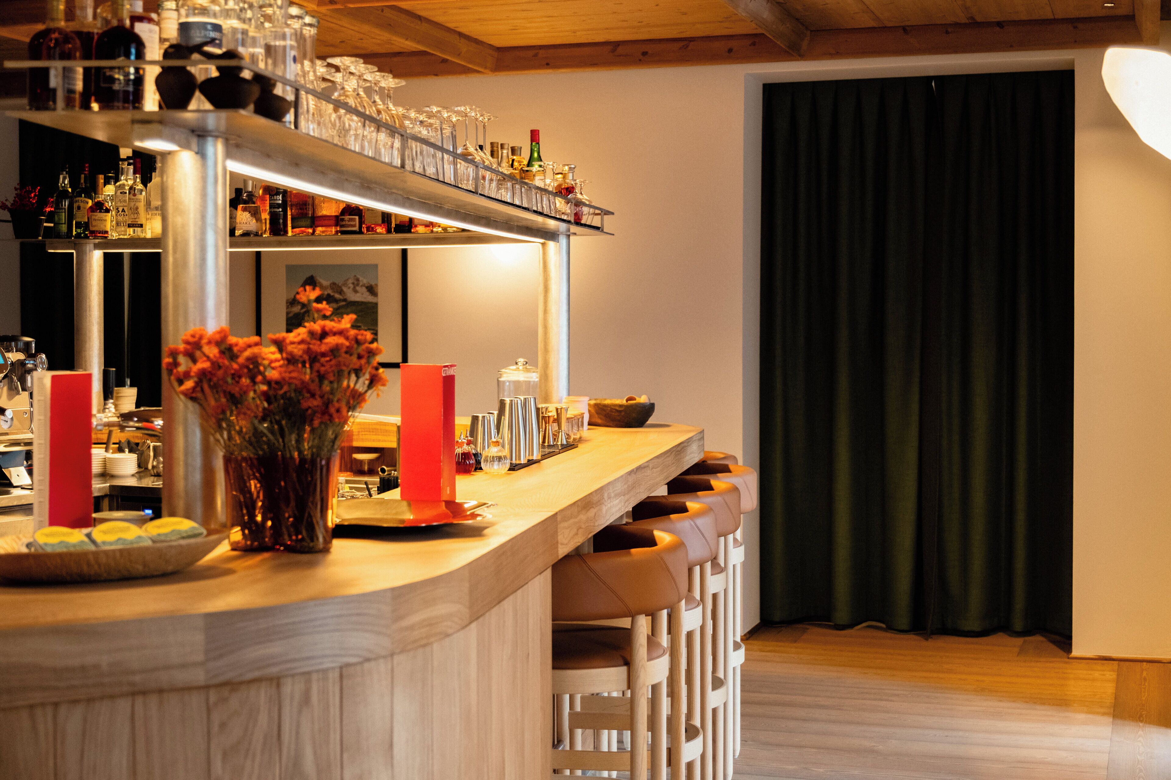 A bar area with a wooden counter, bar stools, a vase with flowers, and various bottles and glasses.