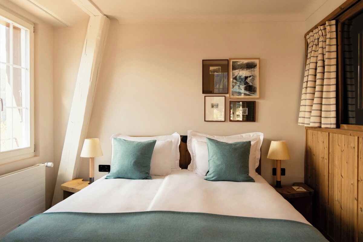 Une chambre à coucher avec un lit, deux tables de chevet avec des lampes, une fenêtre avec des rideaux et des œuvres d’art accrochées au mur.