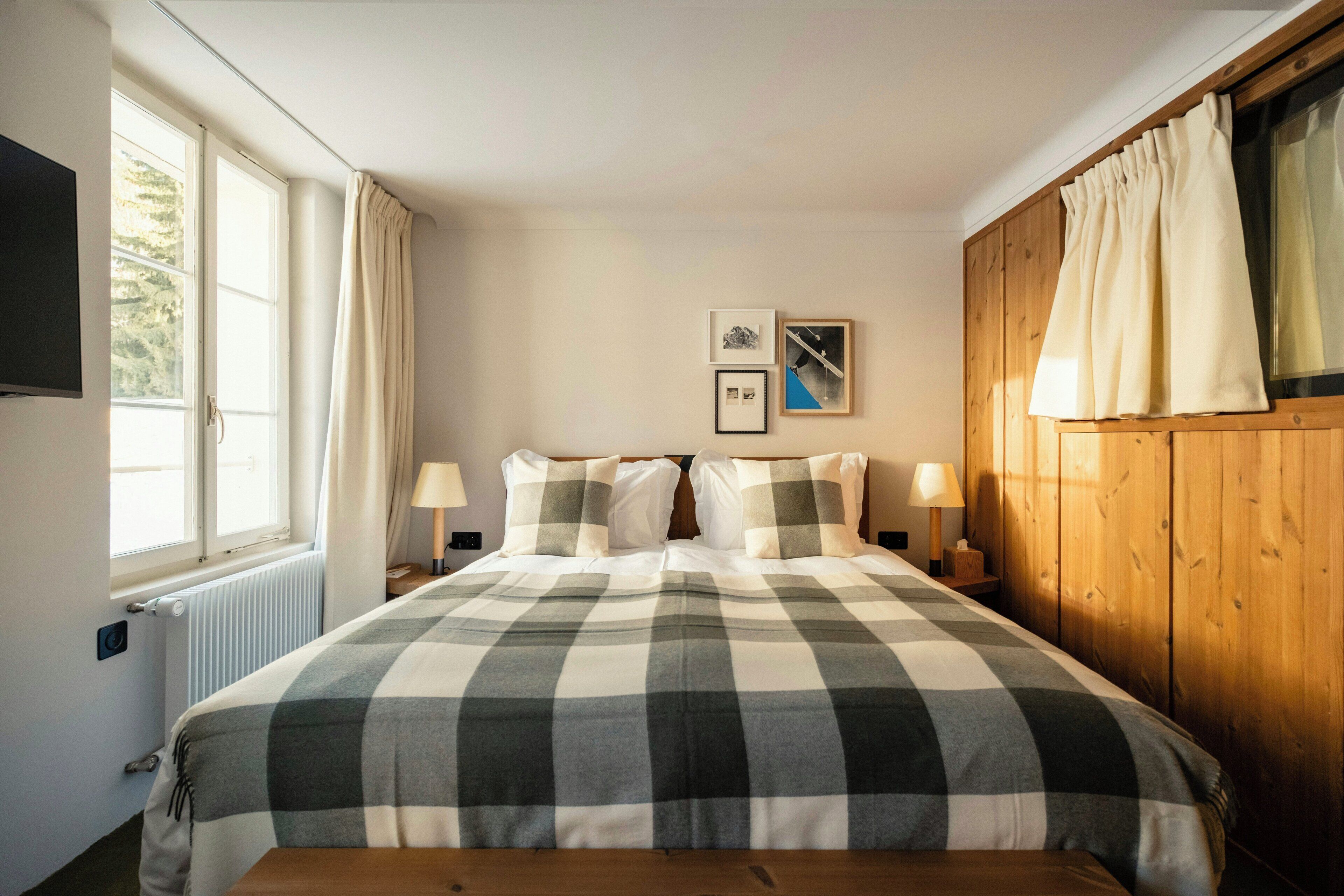 Une pièce comprenant un lit, une fenêtre avec des rideaux, un téléviseur fixé au mur et deux tableaux encadrés.