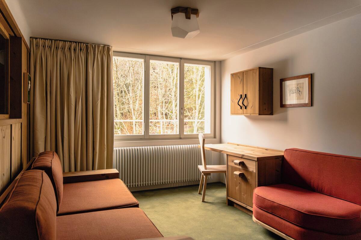 Une pièce comprenant un canapé, un bureau et une chaise. Il y a une fenêtre avec des rideaux, un radiateur et un meuble sur lequel est accroché un tableau au mur.
