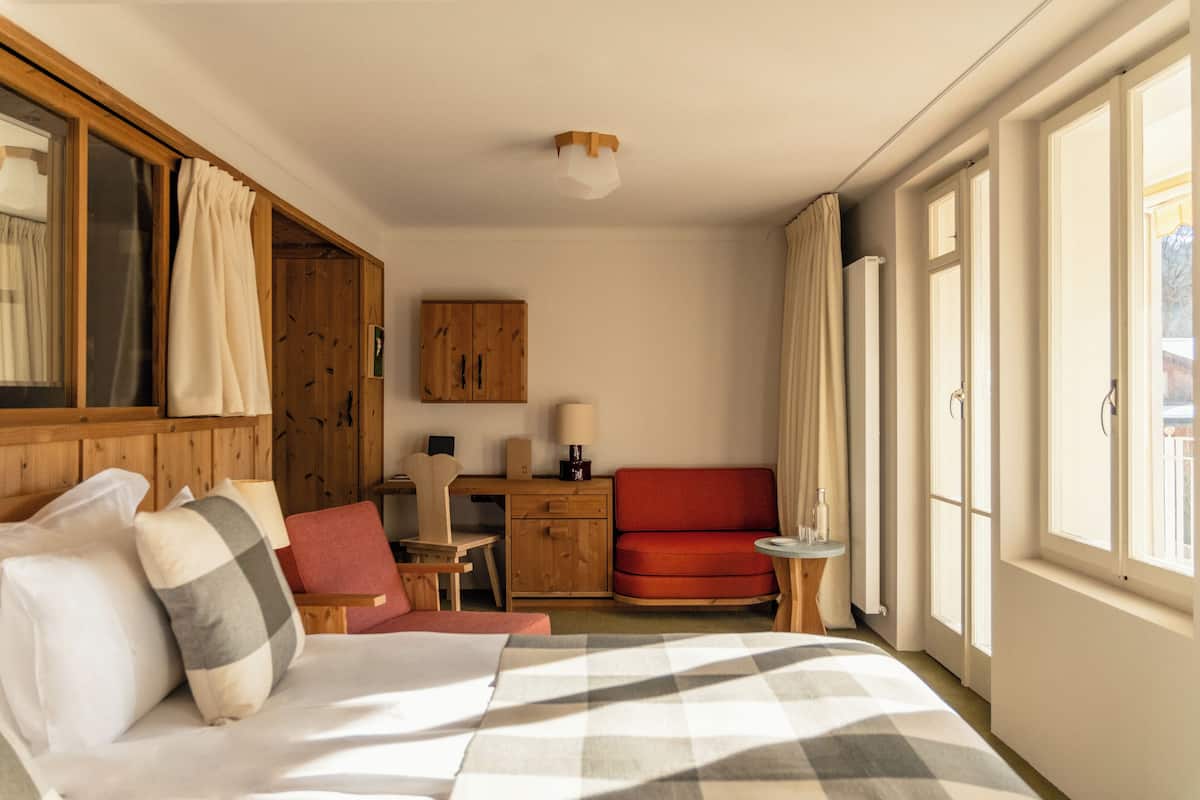 Une chambre d’hôtel avec un lit, un bureau, une chaise rouge, une petite table et une fenêtre avec des rideaux.