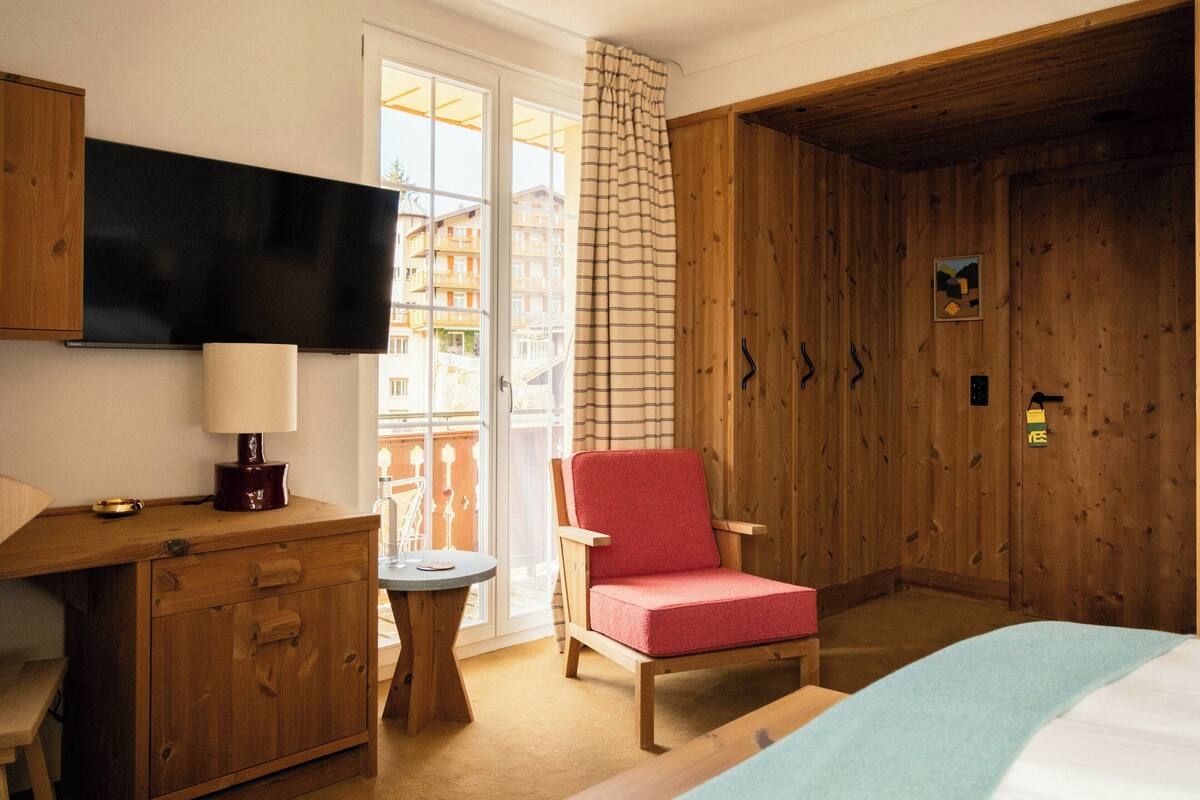 Une pièce avec une armoire en bois, une chaise rouge, un lit, un bureau avec une lampe et un téléviseur fixé au mur.