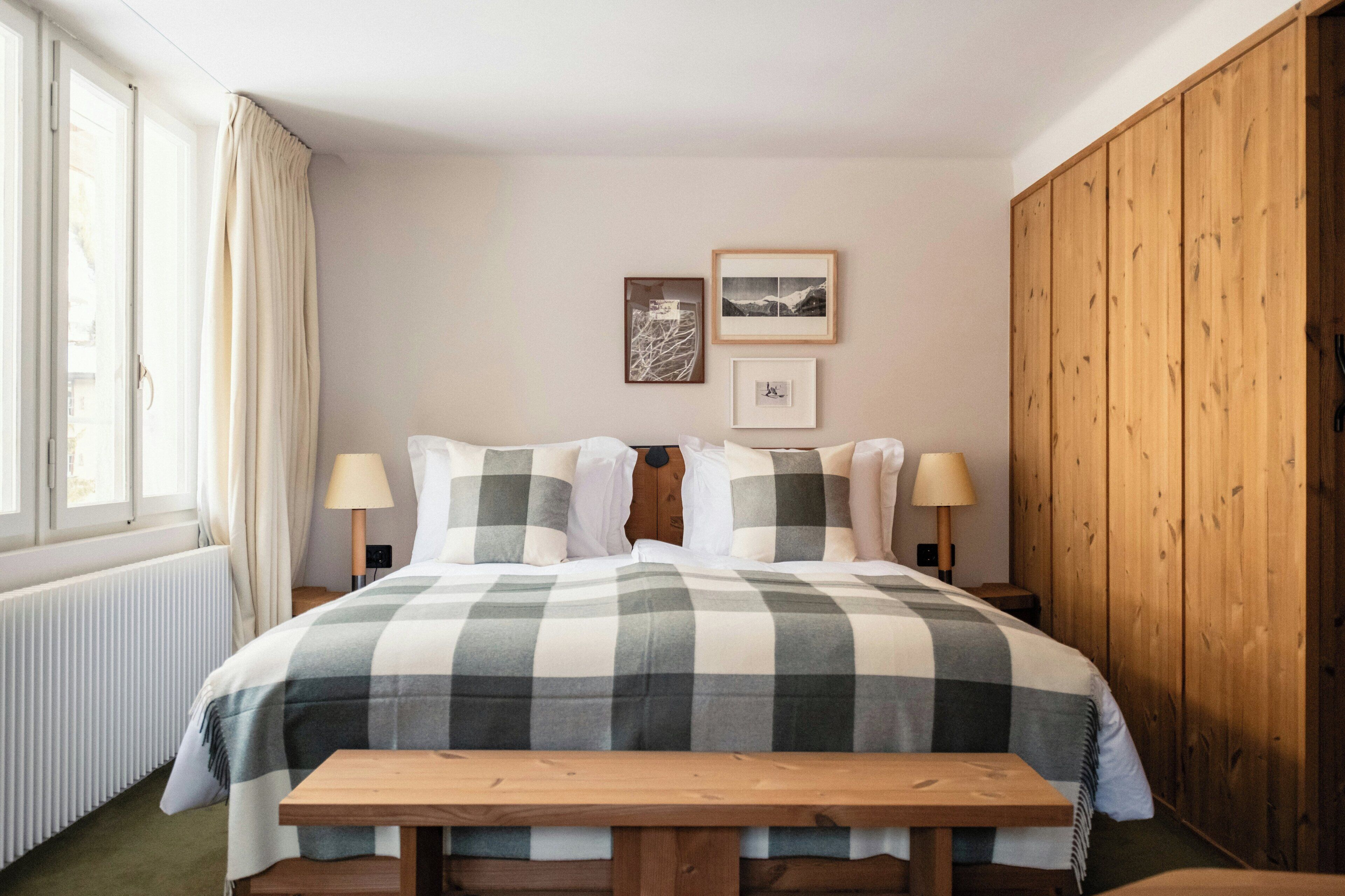 A bedroom with a checkered bedspread, two bedside lamps, a wooden bench, and a window with curtains.
