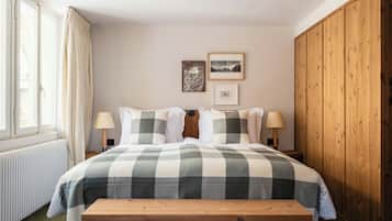 A bedroom with a checkered bedspread, two bedside lamps, a wooden bench, and a window with curtains.
