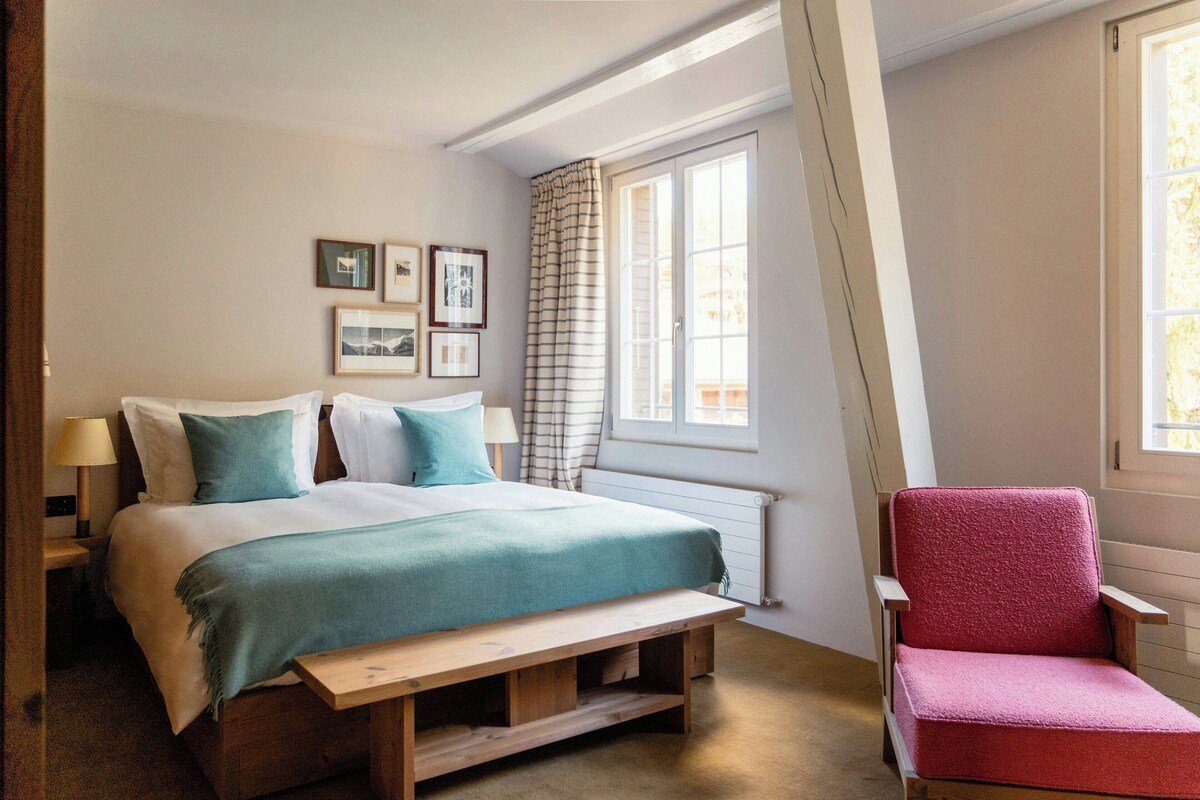 Une chambre à coucher avec un lit, un banc, une chaise, une fenêtre avec des rideaux et des tableaux encadrés accrochés au mur.
