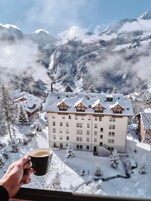 Una mano sosteniendo una taza, con vistas a un edificio cubierto de nieve con múltiples ventanas y un estilo arquitectónico tradicional, frente a un paisaje montañoso.