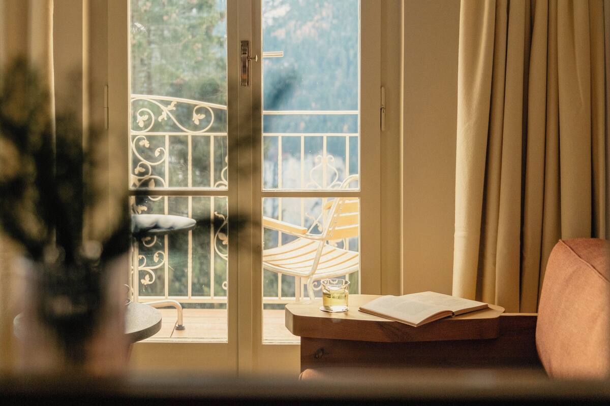 Une pièce avec une fenêtre donnant sur un beau paysage, une chaise, une table avec un livre et un verre, et une plante au premier plan.