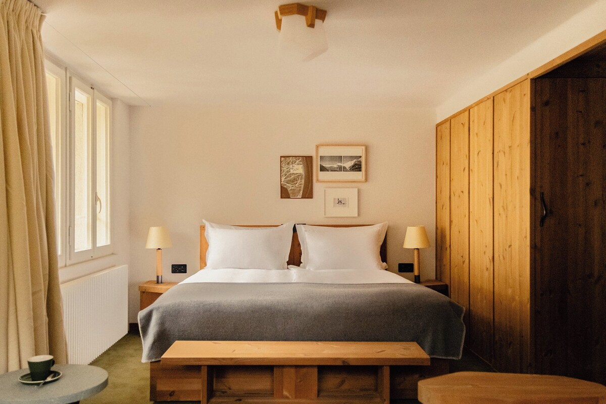 Une chambre à coucher avec un lit, des lampes de chevet, un banc en bois, une petite table avec une tasse, et une fenêtre avec des rideaux.