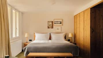 A bedroom with a bed, bedside lamps, a wooden bench, a small table with a cup, and a window with curtains.