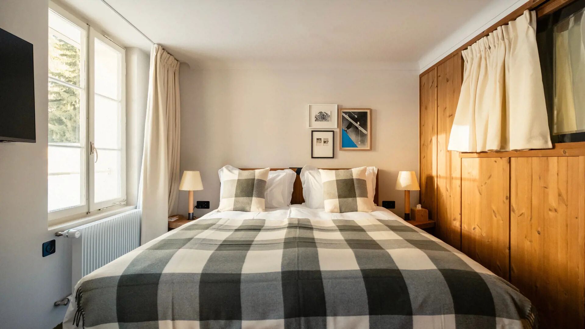 A hotel room with a large bed, two bedside tables with lamps, a window with curtains, and a wooden wall panel.
