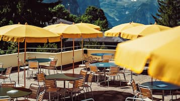 Terrasse mit gelben Sonnenschirmen, Tischen und Stühlen vor einer Bergkulisse.