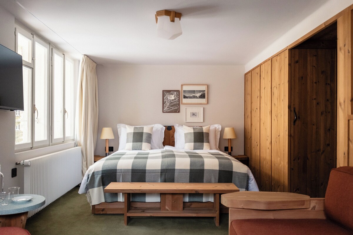 Une chambre à coucher avec un grand lit, deux lampes de chevet, un banc en bois, une télévision et une fenêtre avec des rideaux.