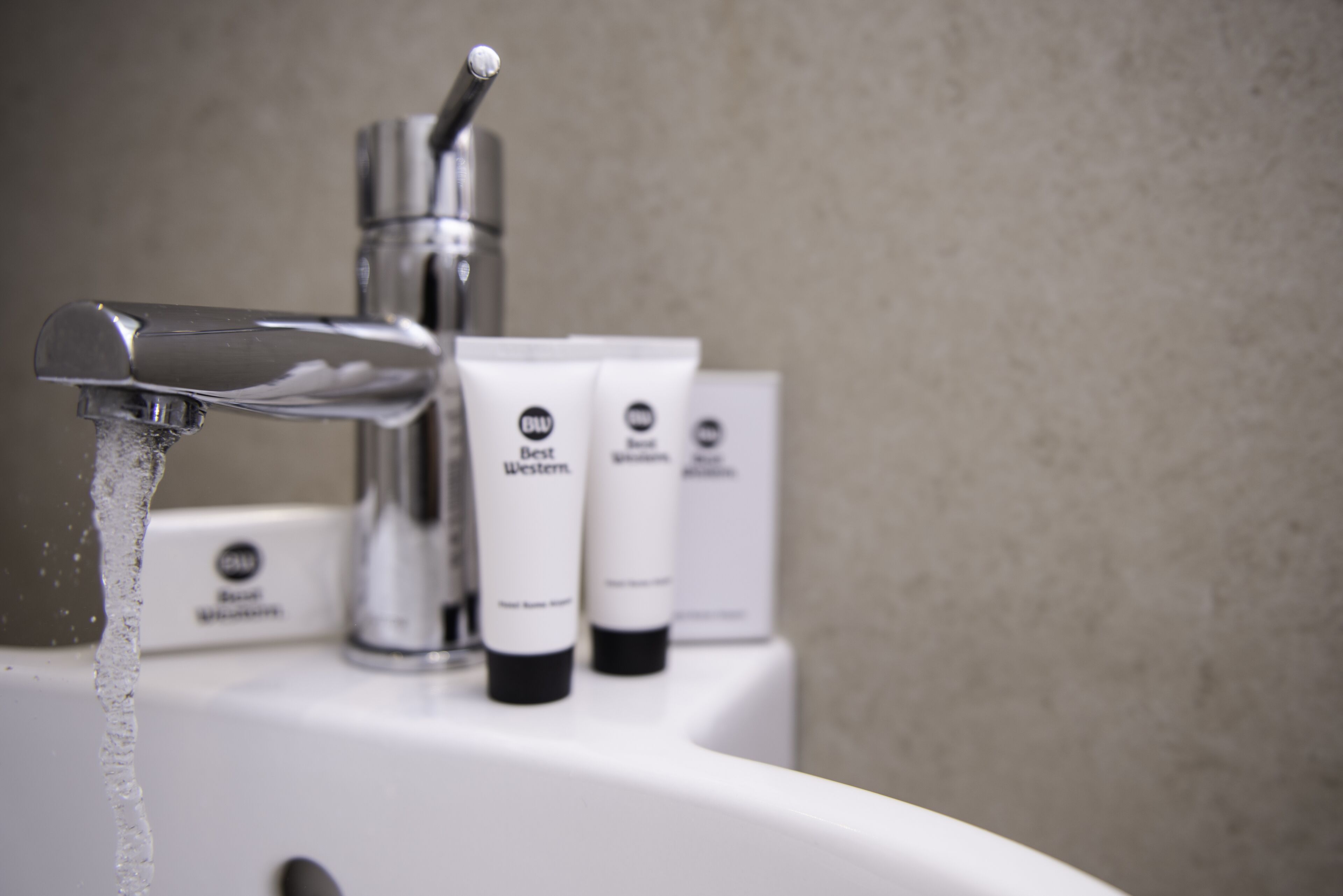 Un lavamanos de baño con agua corriendo, un grifo moderno y tres botellas blancas con la etiqueta 'Best Western'.