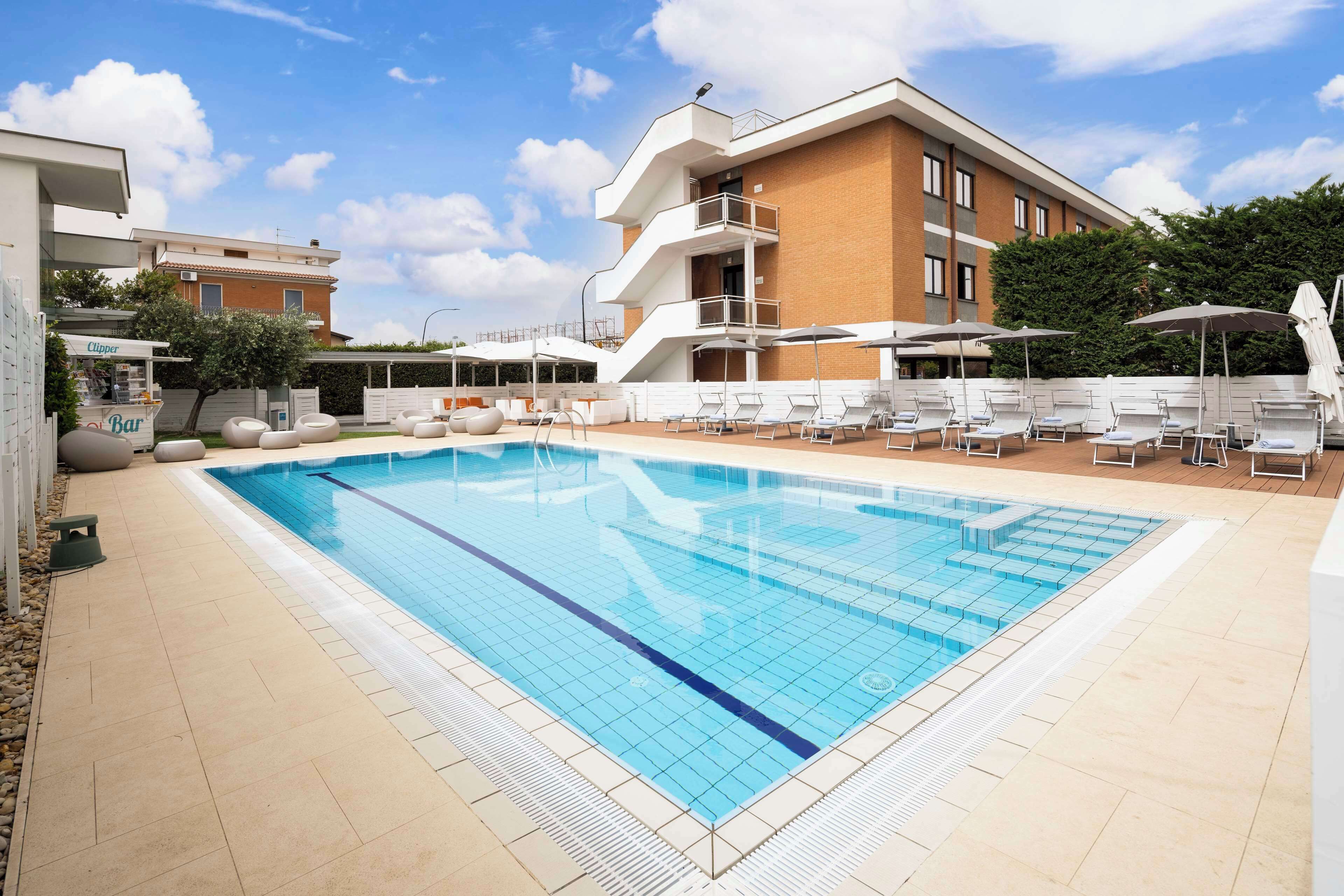 Une piscine avec des chaises longues et des parasols, un bar et un immeuble à plusieurs étages doté de balcons.