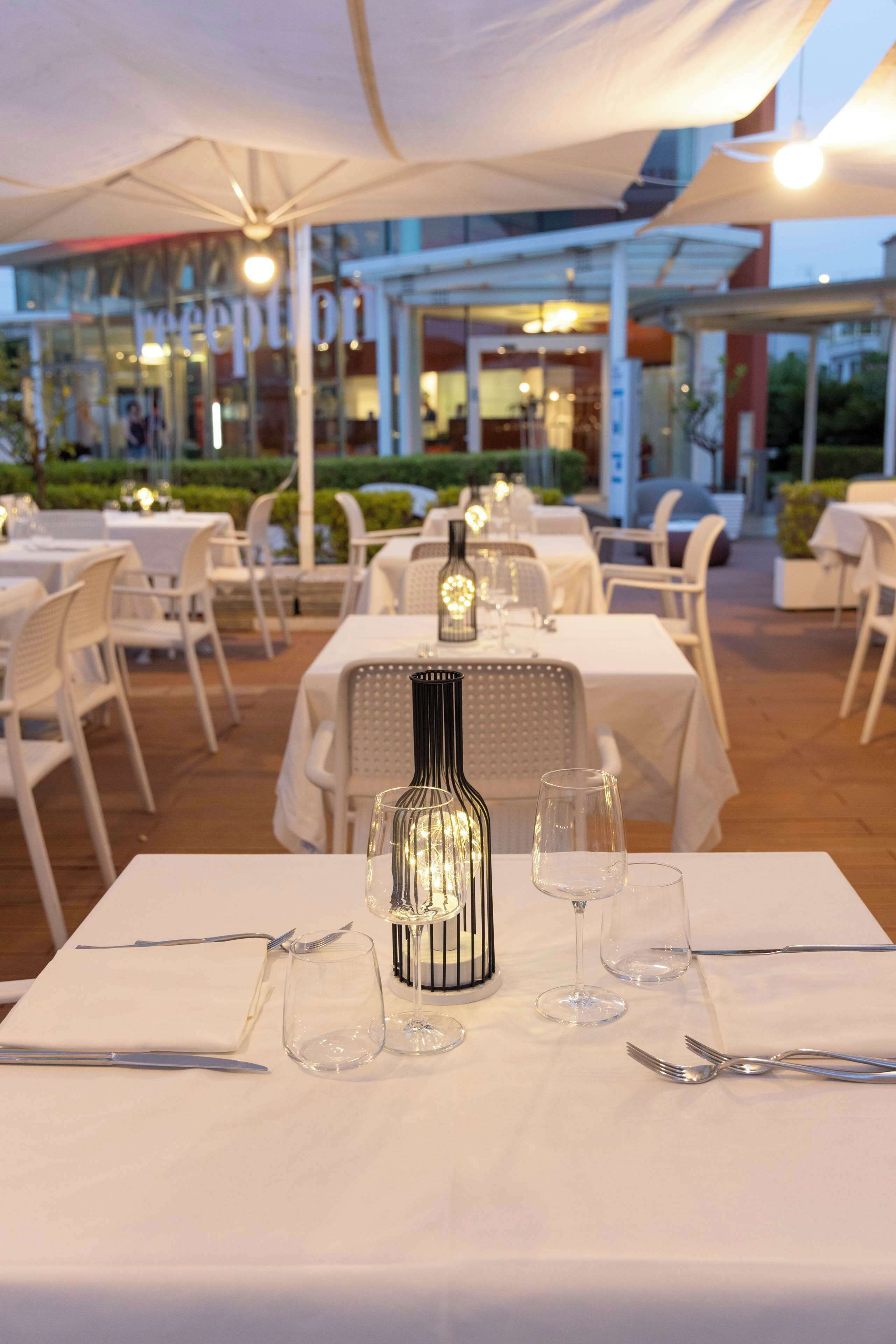 A dining area with tables set for two, featuring white tablecloths, wine glasses, and cutlery.