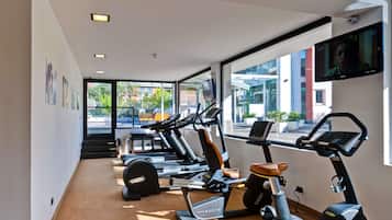 Un gimnasio con varias máquinas de cardio, un televisor en la pared y grandes ventanales que ofrecen vistas al exterior.
