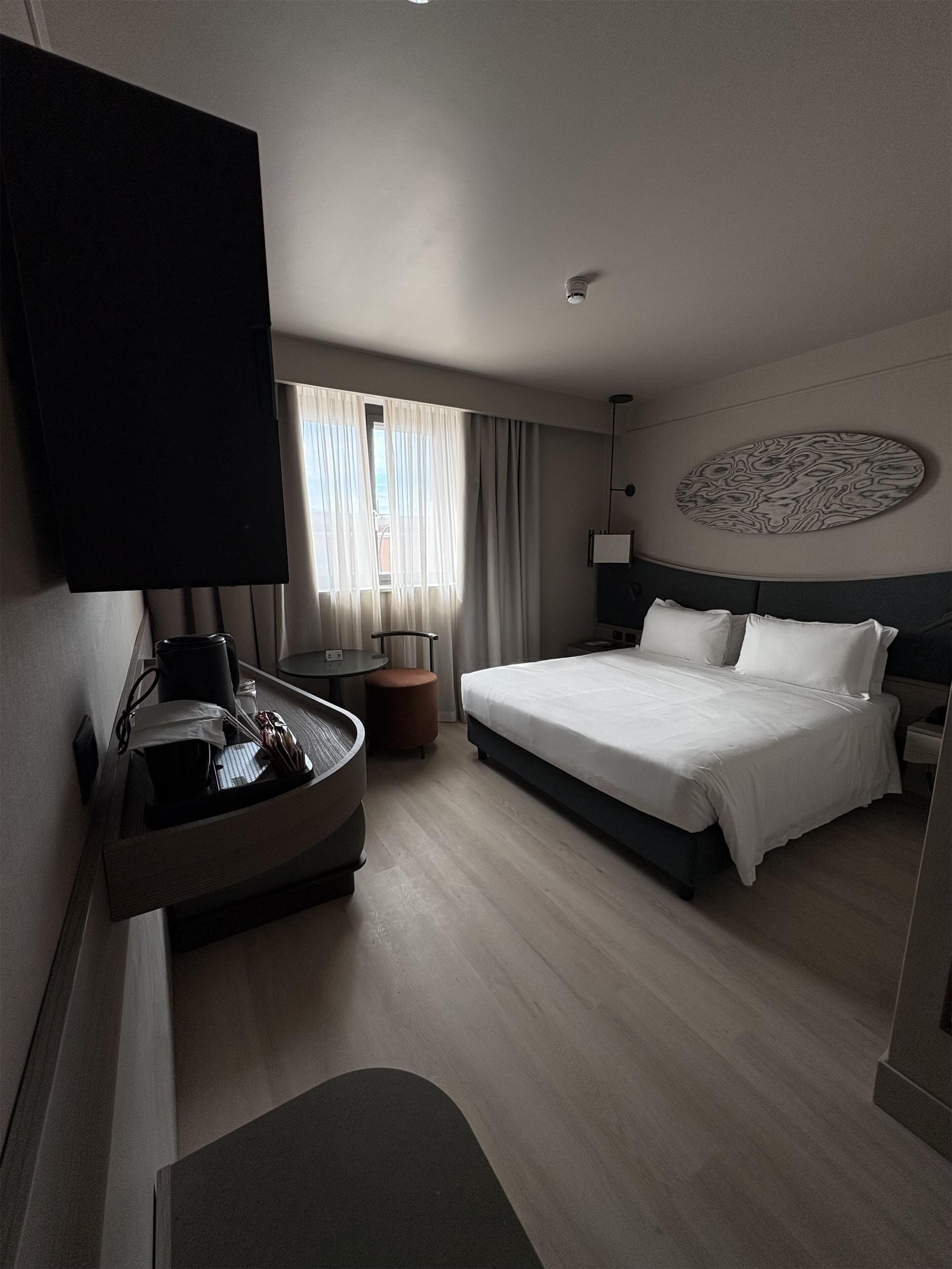 A hotel room with a bed, a desk with a chair, a television, a wall-mounted artwork, and a window with curtains.