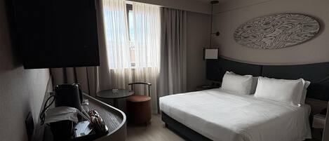 A hotel room with a bed, a desk with a chair, a television, a wall-mounted artwork, and a window with curtains.