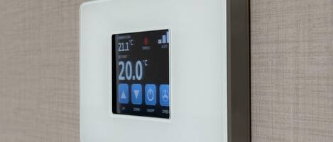 Termostato empotrado en la pared que muestra una temperatura de 21,1°C y un ajuste de 20,0°C, con cuatro botones de control etiquetados como SUBIR, BAJAR, ENCENDIDO/APAGADO y VELOCIDAD.