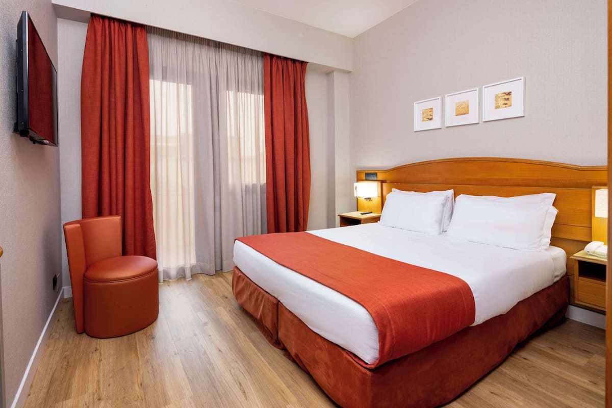 A hotel room with a large bed, a chair, a TV, and a nightstand with a lamp.