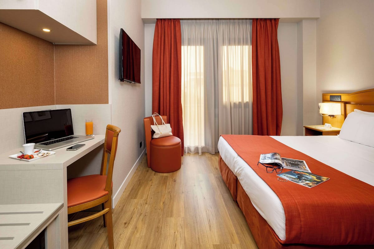 A hotel room with a desk, a bed, a chair, a TV, and a window with curtains.