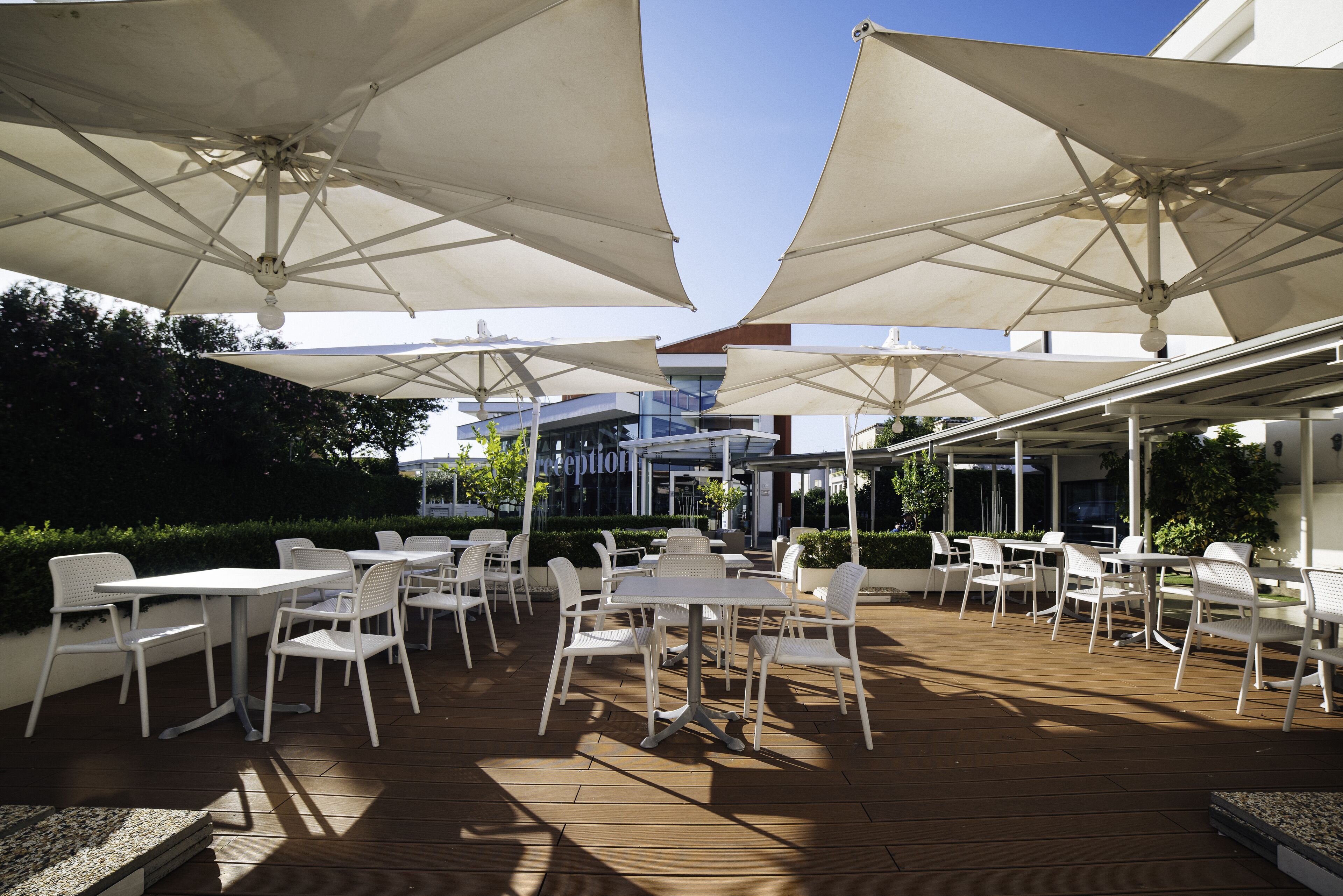 An outdoor patio with white tables and chairs under large umbrellas, surrounded by greenery and a building in the background.