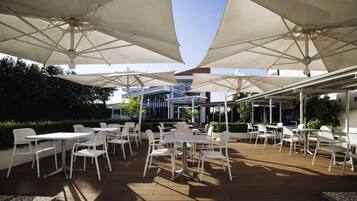 An outdoor patio with white tables and chairs under large umbrellas, surrounded by greenery and a building in the background.