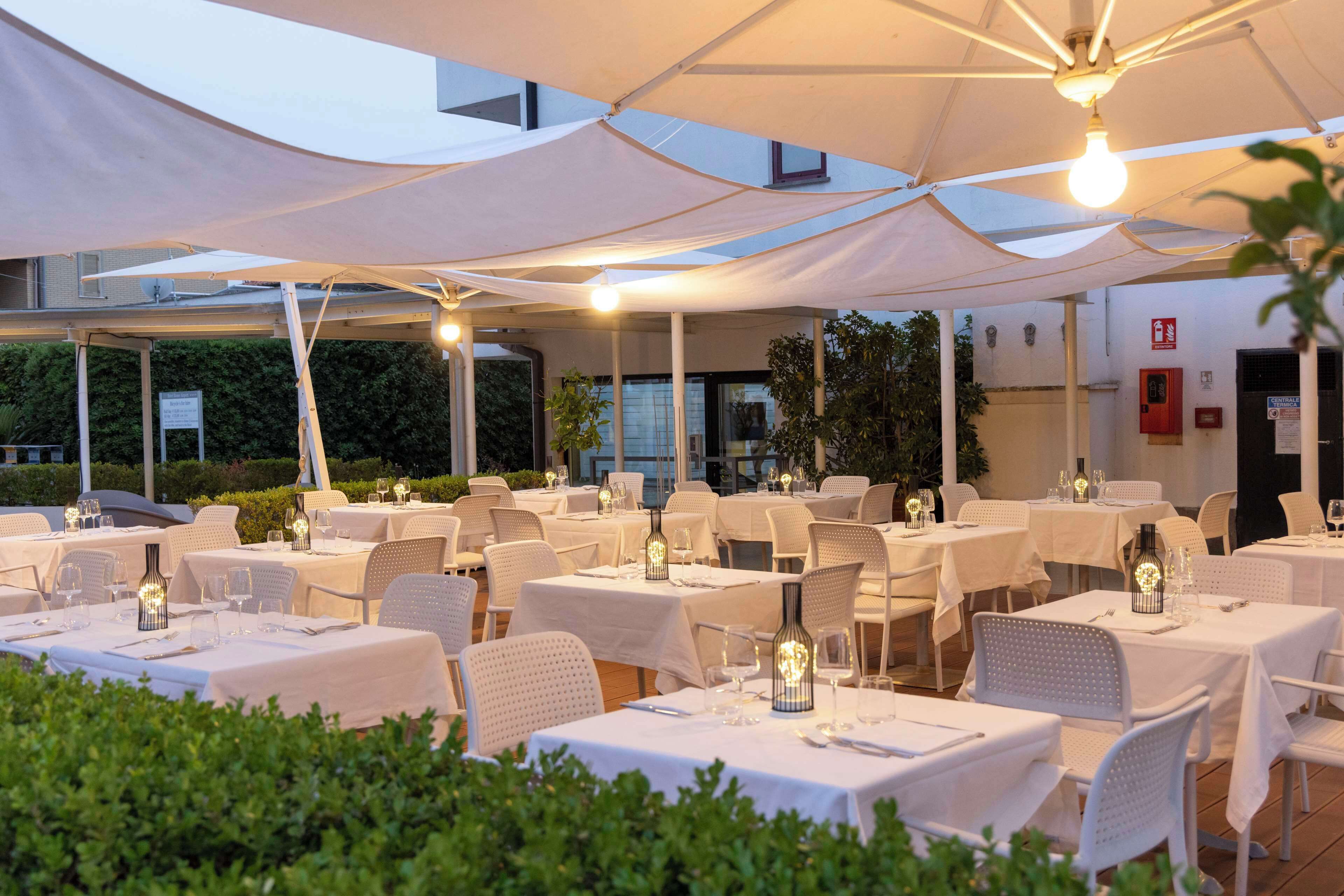 An outdoor dining area with tables set for two, covered by white canopies and lit by hanging light fixtures.