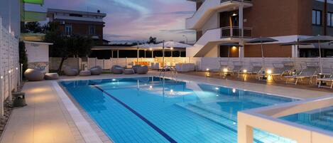 Una piscina con sillas de descanso y sombrillas, rodeada de edificios con balcones y terrazas.