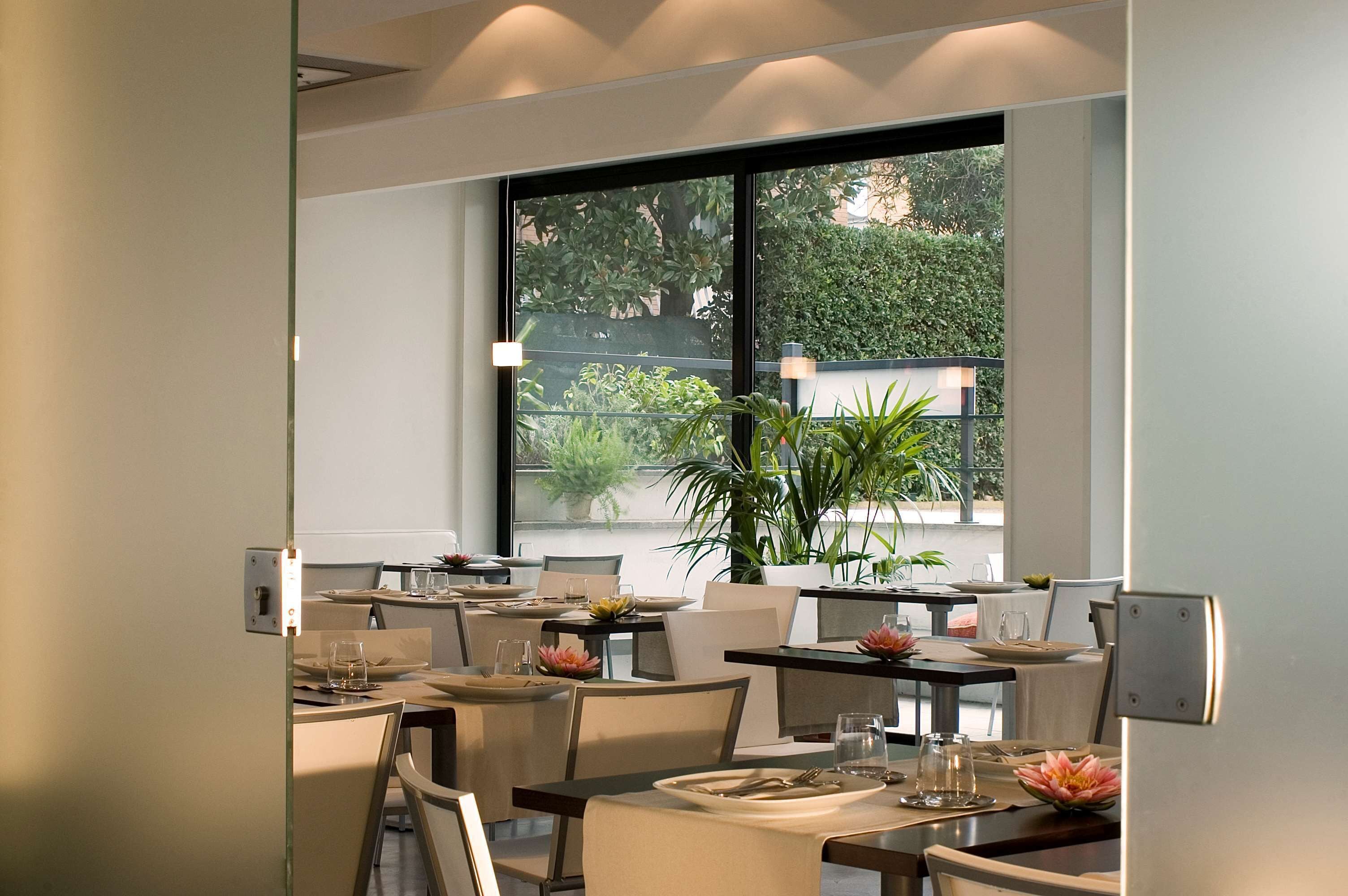 A modern dining area with tables set for two, large windows with a view of greenery, and potted plants on the windowsill.