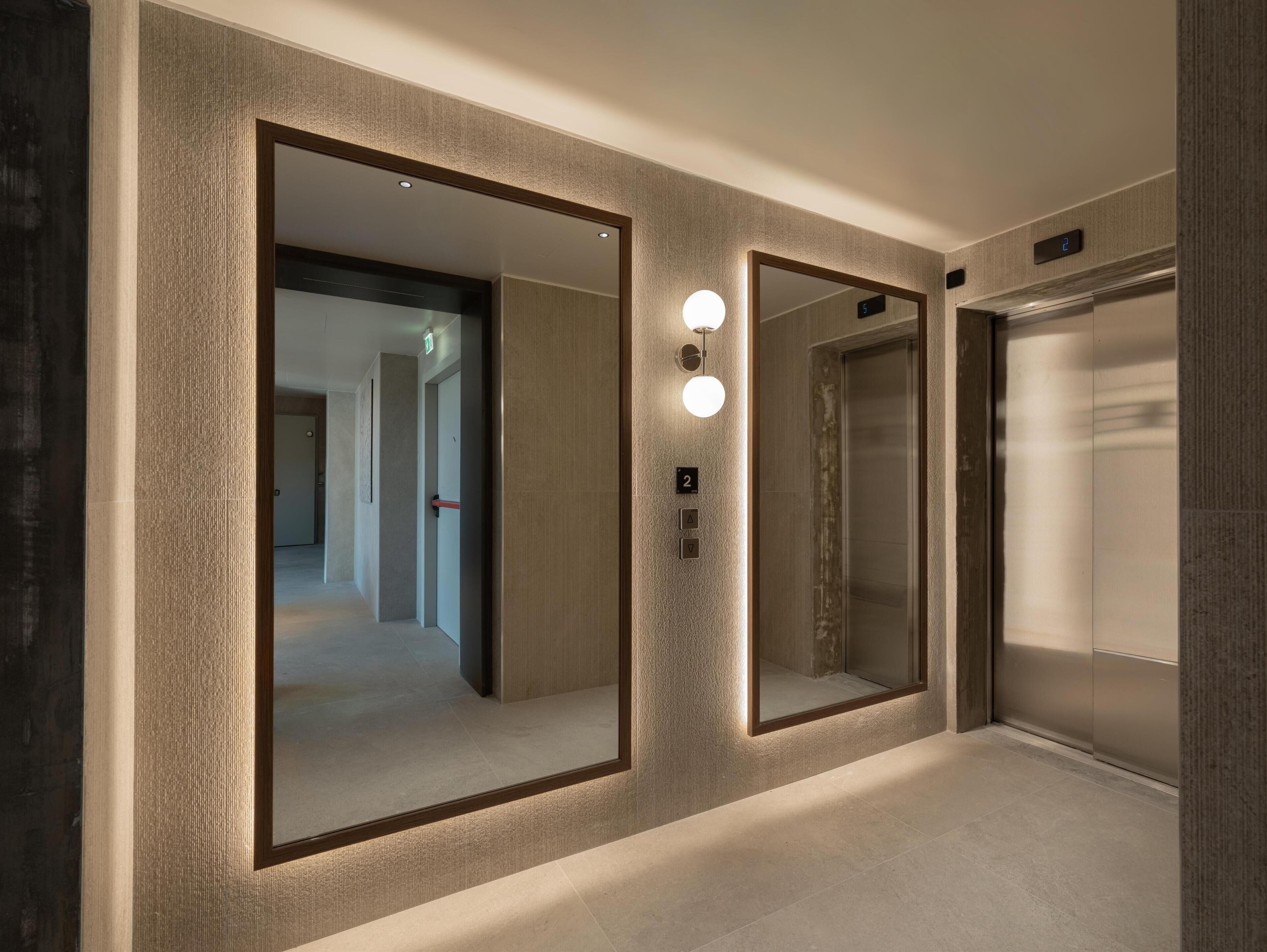 Un couloir avec deux ascenseurs, un miroir et une porte portant un panneau indiquant la sortie de secours.