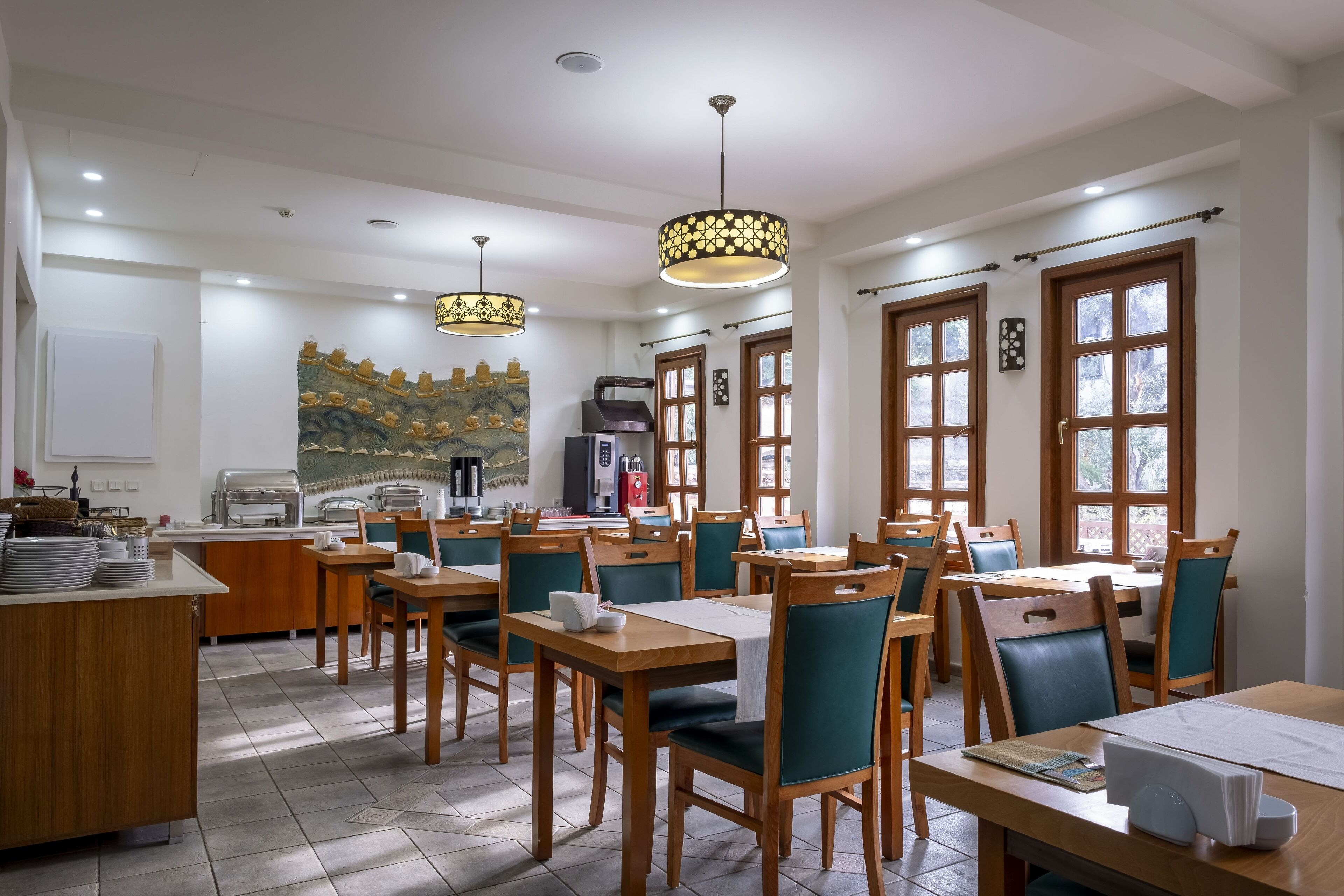 A dining area with wooden tables and chairs, a kitchen in the background, and large windows.