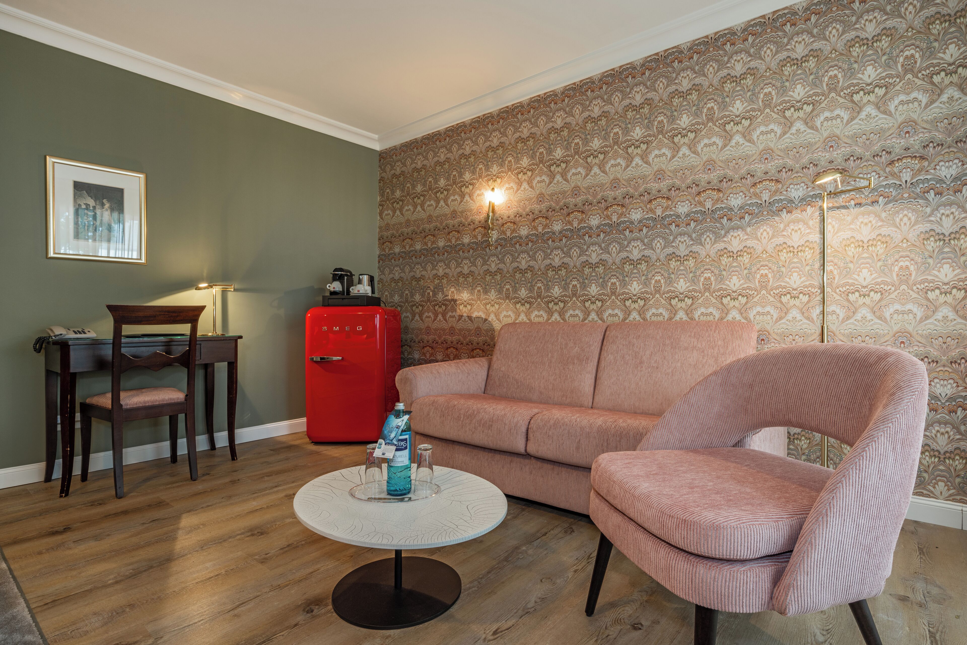 Una sala de estar con piso de madera, un refrigerador rojo, un sofá rosa, una mesa de centro redonda y un cuadro en la pared.