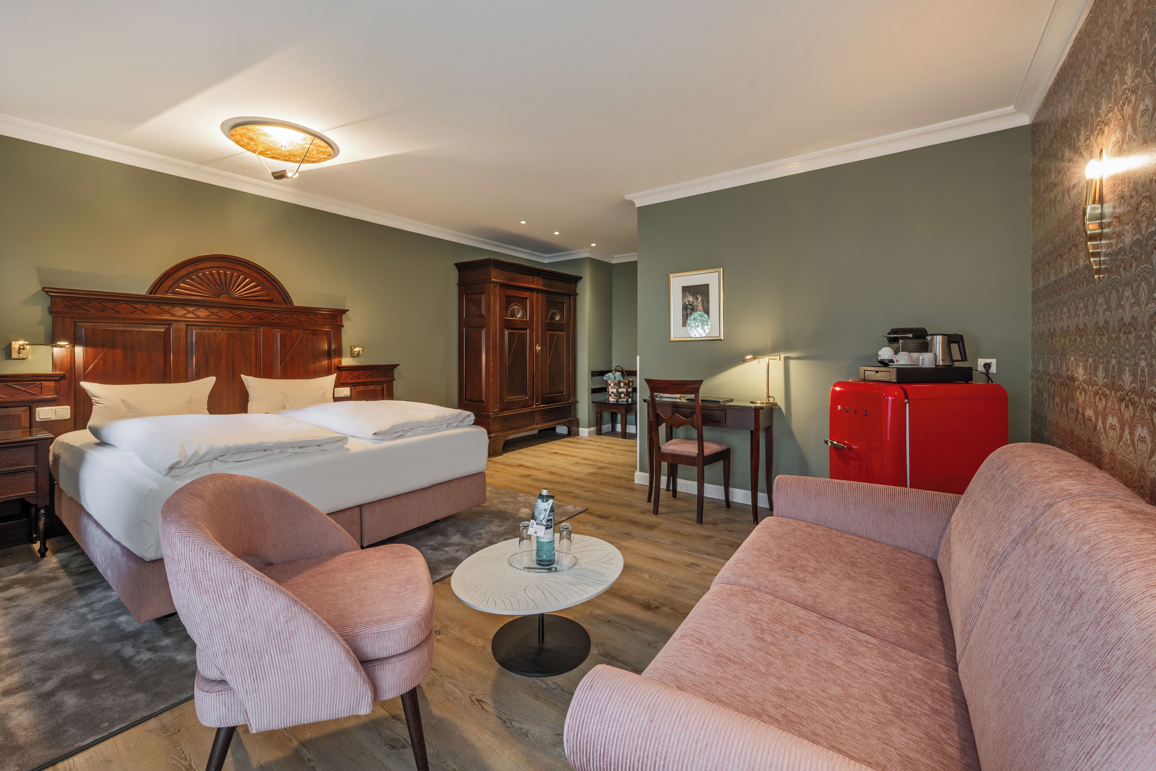 A hotel room with a large bed, two armchairs, a small table, a red refrigerator, and a wooden cabinet.