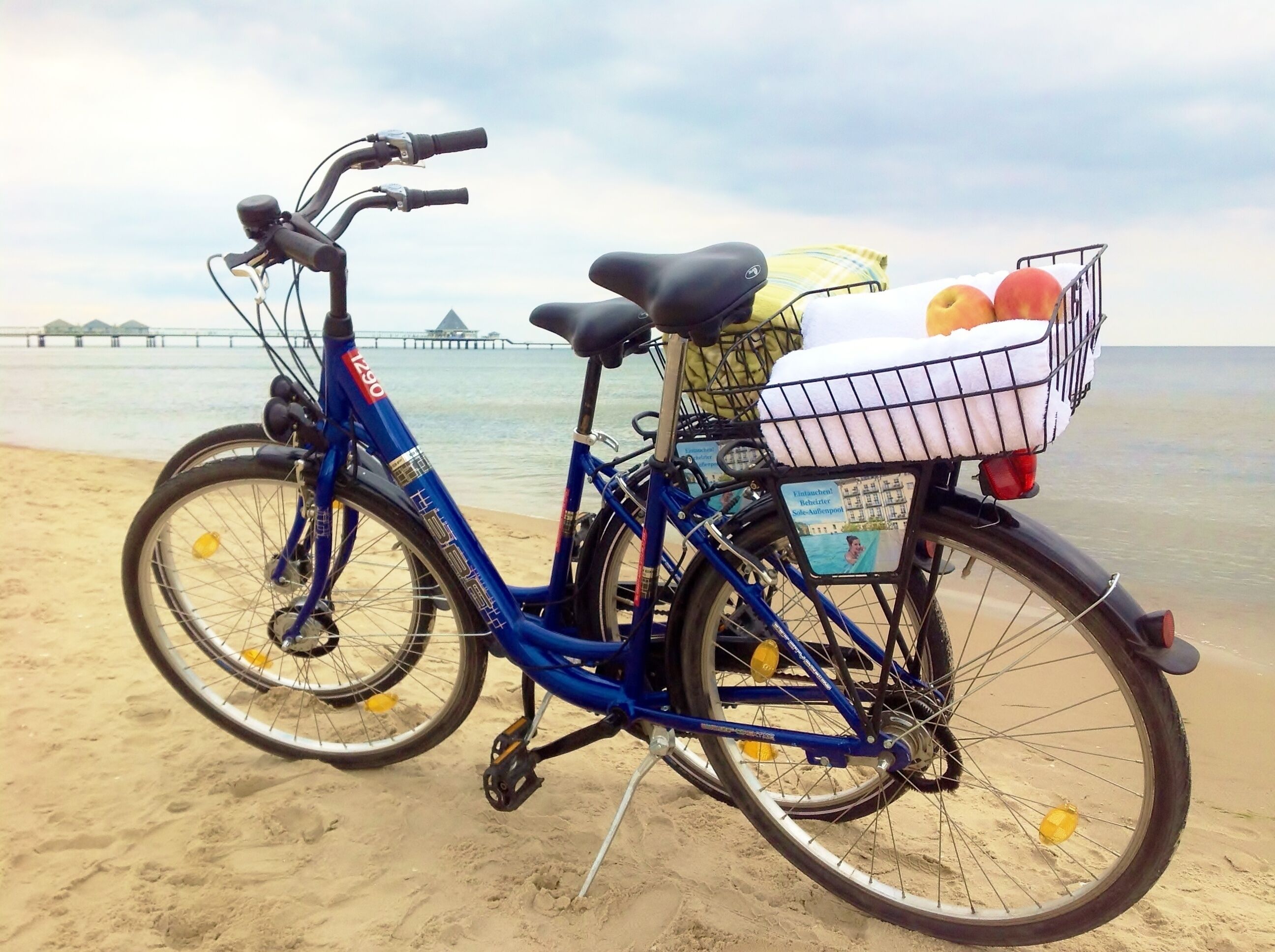 A blue bicycle with a basket on the back, parked on a sandy beach with a body of water and a pier in the background.
