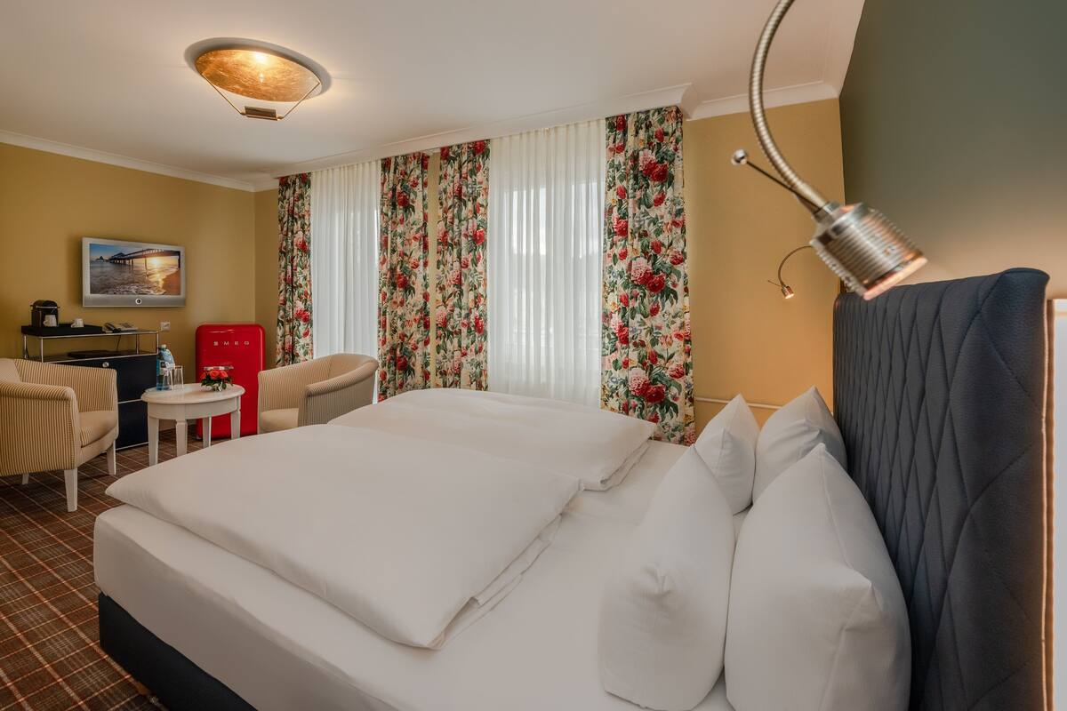 Ein Hotelzimmer mit einem großen Bett, einem Schreibtisch mit Stuhl, einem kleinen Tisch und blumigen Vorhängen.