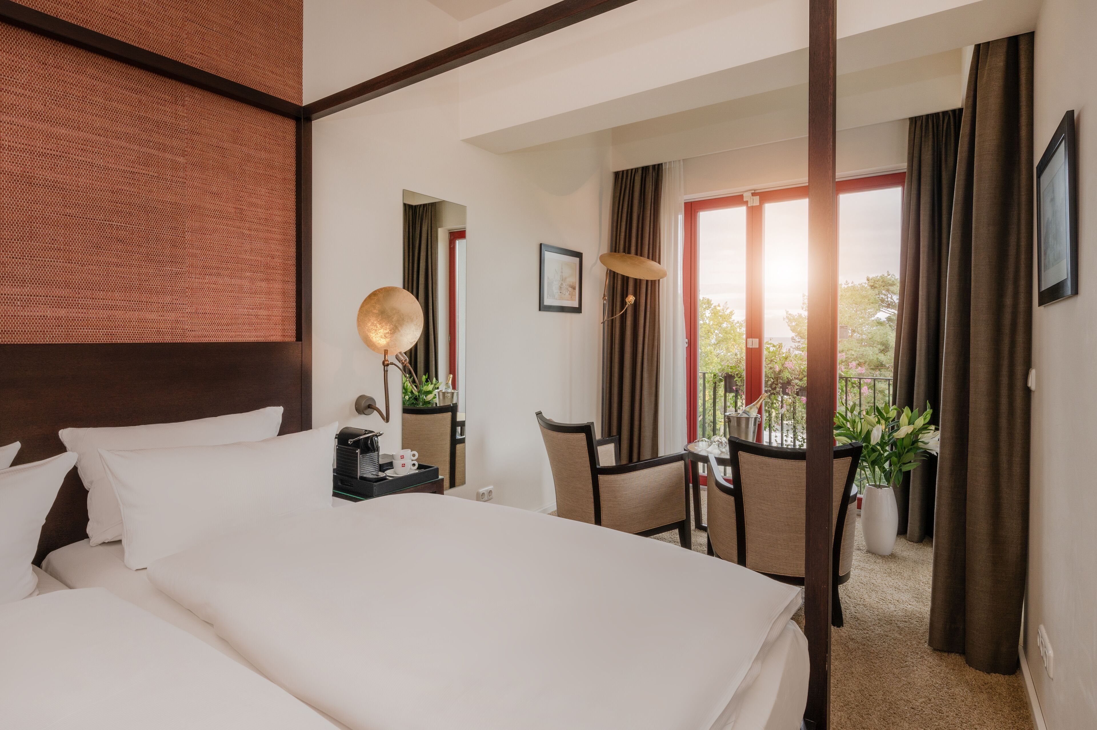 Ein Hotelzimmer mit einem großen Bett, einem Schreibtisch mit Stuhl, einem kleinen Tisch mit Pflanze und Blick ins Freie durch eine Glastür.