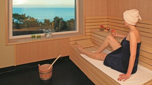 A sauna room with a wooden bench, a window with a view of the sea, and a bucket with a wooden spoon.