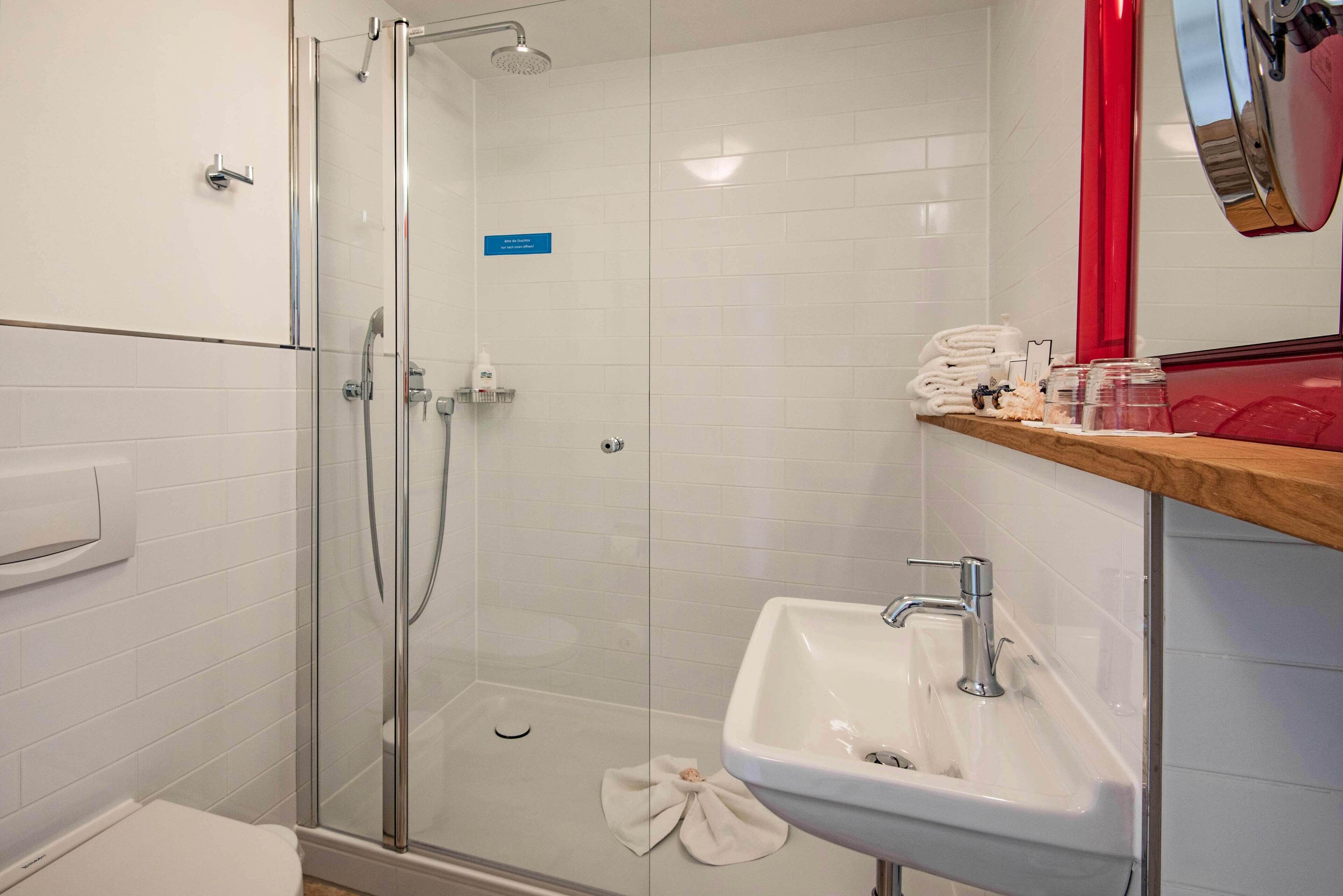 Une salle de bain moderne avec une douche à parois de verre, un lavabo blanc et une étagère en bois où sont rangés les articles de toilette.
