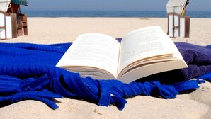 An open book on a blue blanket on a sandy beach with two beach chairs and a small hut in the background.
